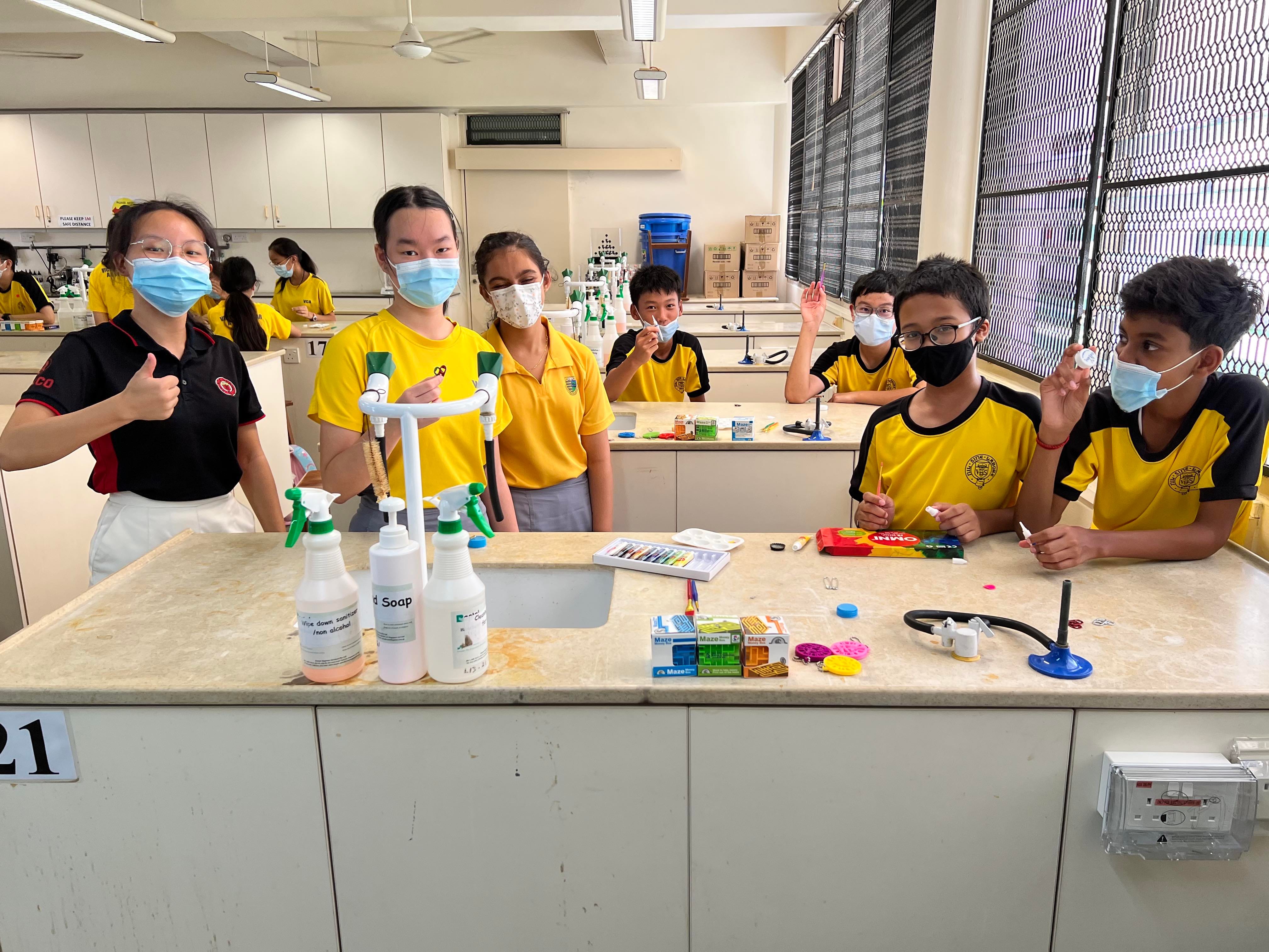 Students in yellow uniforms and face masks pose at a school science lab bench with spray bottles and supplies.